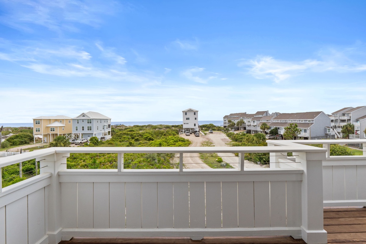 Elevated deck overlooks coastal landscape and neighboring beach properties, providing a serene view of dunes, sea vegetation, and blue sky in this vacation rental setting.