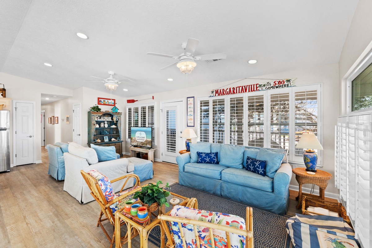 Relax in your bright coastal living room with comfortable seating, ceiling fans, and charming beach-themed decor that creates the perfect vacation atmosphere.
