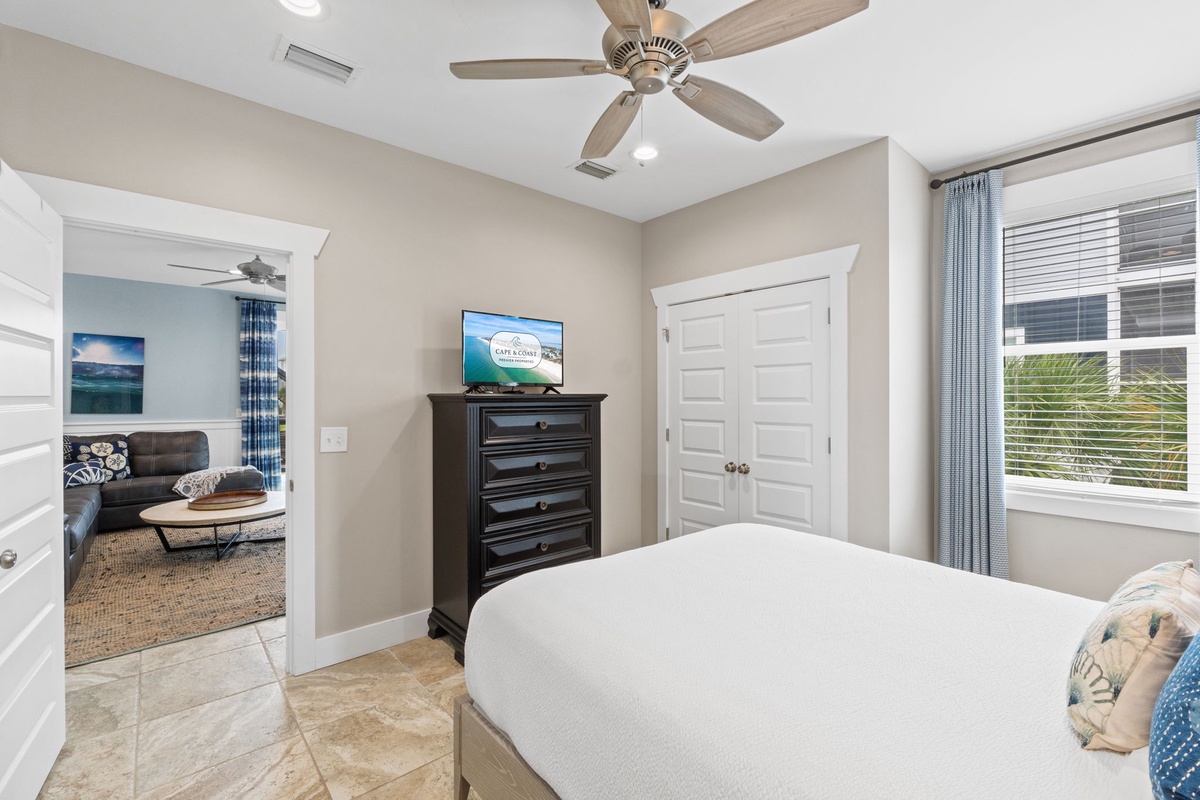 Your peaceful retreat features crisp linens, a ceiling fan to keep you cool, and natural light streaming through the window, with a cozy living area just beyond the doorway.