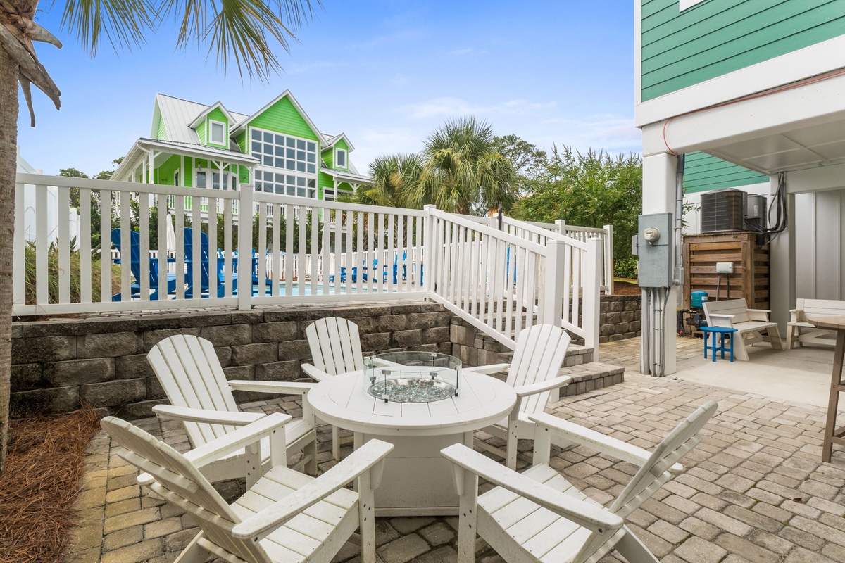 Relax in white Adirondack chairs around a fire pit on your private patio, with views of the vibrant green vacation home and pool beyond.