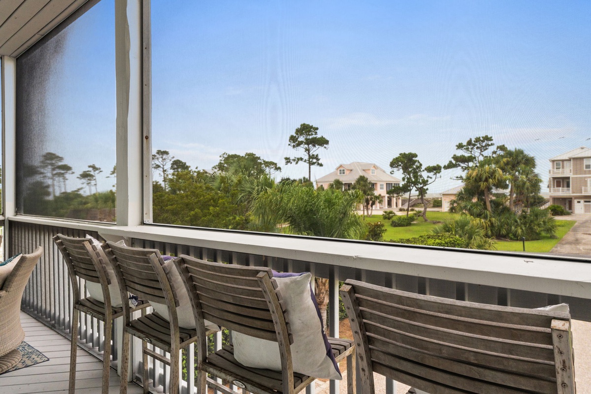 Screened balcony with seating overlooking a peaceful neighborhood of lush greenery, native pines, and coastal homes under clear blue skies.