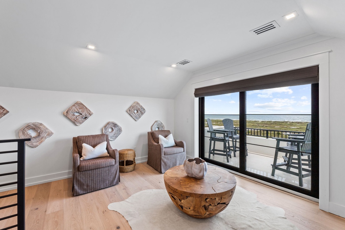 A serene seating area with comfortable chairs, natural wood accents, and coastal decor overlooking a private balcony with sweeping views of marshland and water beyond.
