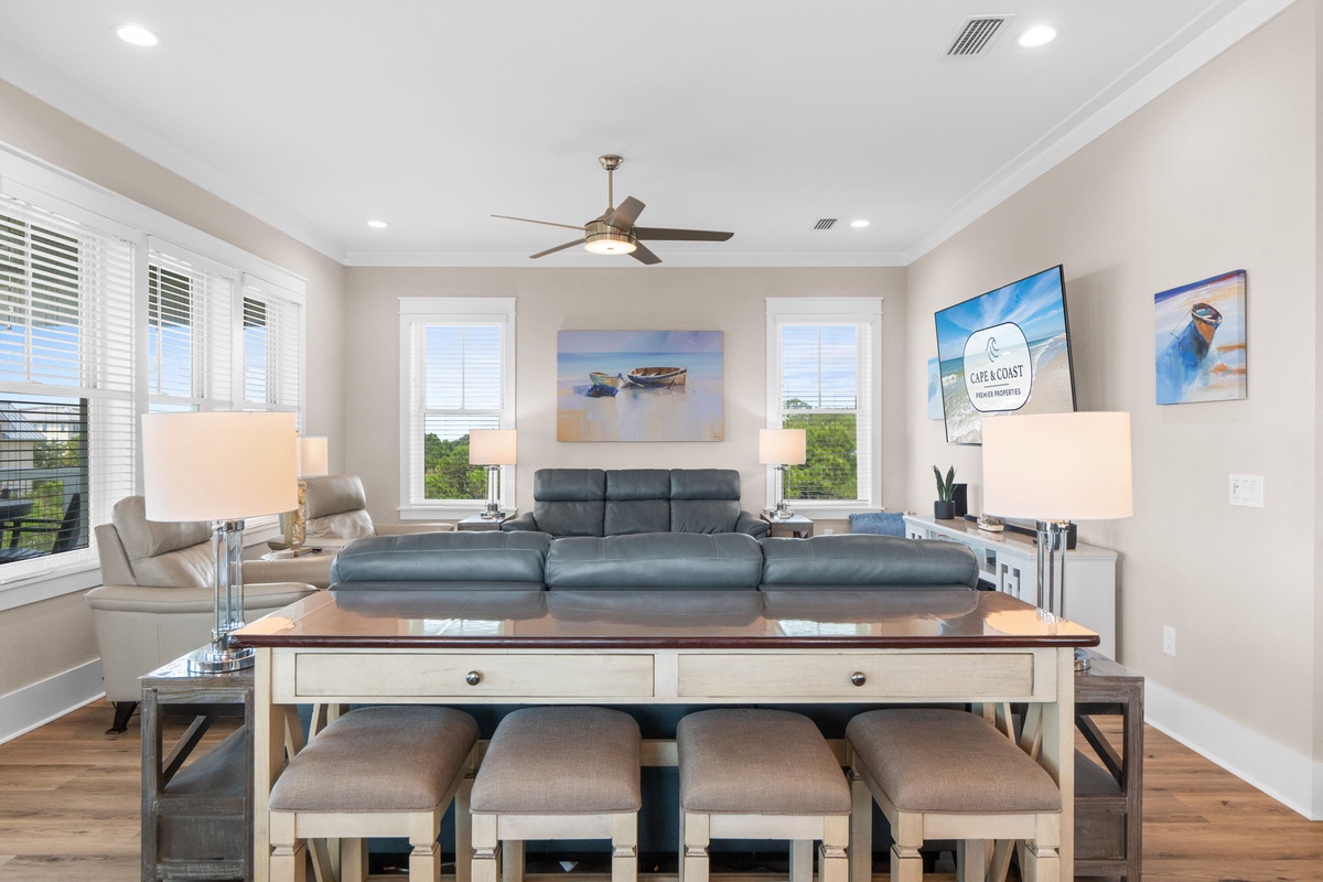 Bright, open living space with vaulted ceilings, coastal artwork, and windows framing greenery. Features a versatile console table, plush sofa, and cozy armchair—your perfect retreat awaits.