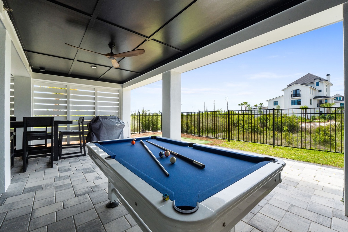 Covered outdoor game area with a billiards table, bar seating, and ceiling fan for your entertainment, overlooking a green lawn and neighborhood backdrop.