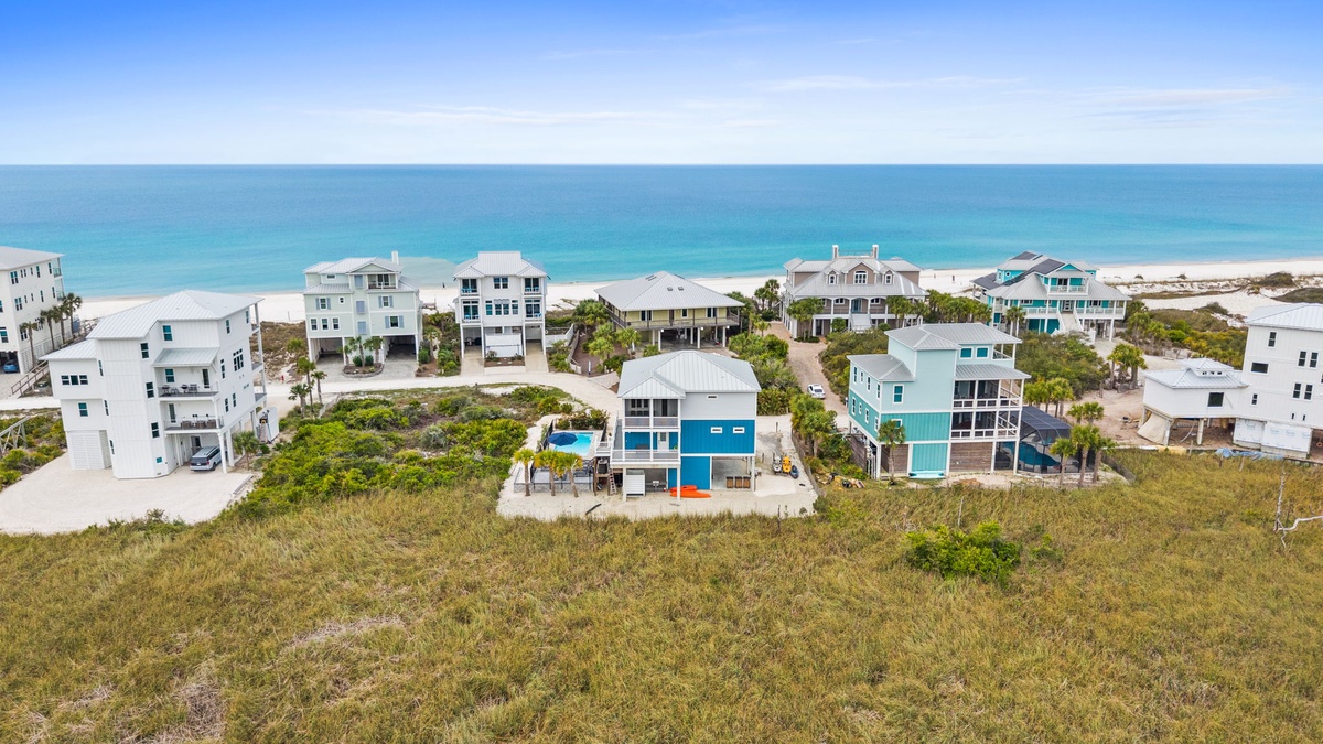 Aerial view shows colorful beach homes nestled between natural dunes and pristine coastline, with crystal-clear waters stretching to the horizon.