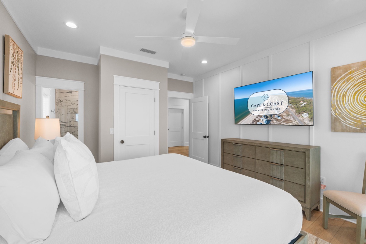 Sink into your plush bed and unwind in a bright, coastal-chic bedroom with custom art, a mounted TV, and ceiling fan—your perfect retreat after a day exploring.