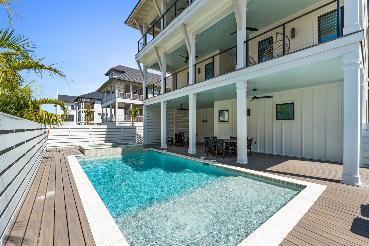 A modern coastal villa featuring a private pool surrounded by wooden decking, white columns, covered outdoor lounging areas, and palm trees—an inviting setting in a residential resort community.