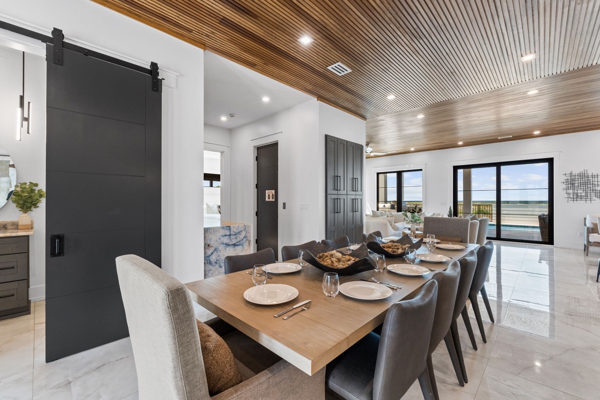 Picture yourself gathering with loved ones at this stunning dining table, where a striking wood-slatted ceiling and elegant barn door create a sophisticated coastal retreat. Your view stretches to the water through oversized windows, blending modern luxur