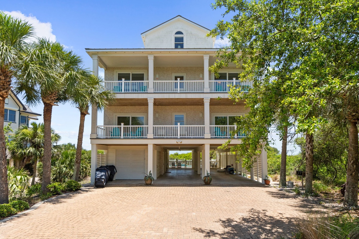 A three-story coastal vacation home featuring spacious covered balconies, elevated design with parking below, and lush tropical landscaping including mature palm trees and greenery.