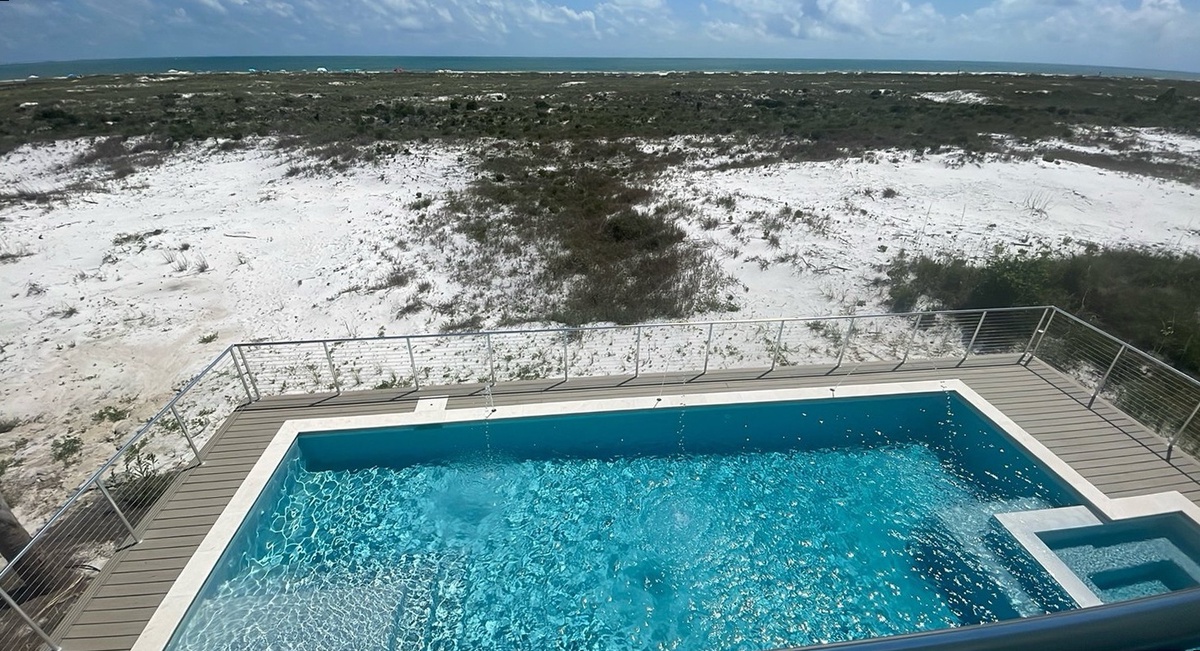Elevated pool deck overlooking white sand dunes and turquoise ocean waters, offering a serene coastal setting for the property's outdoor amenities.