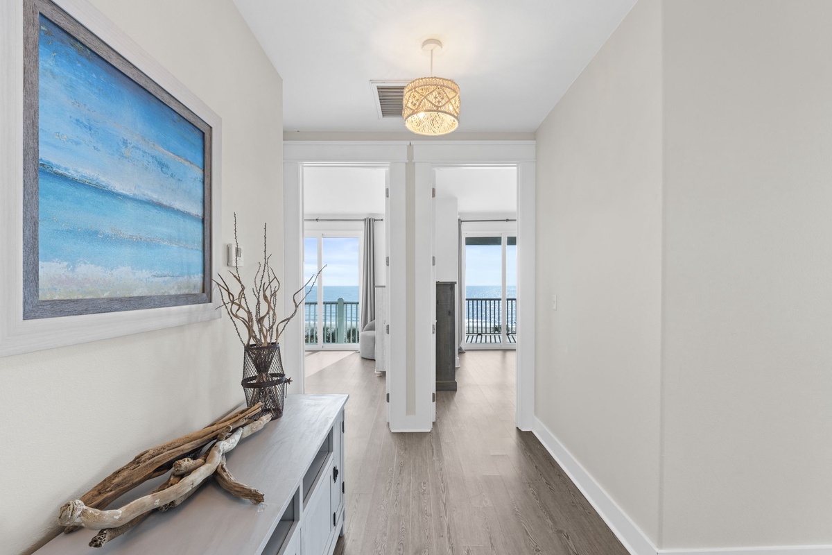 Step into your coastal hallway where ocean artwork and warm lighting guide you toward stunning sea views through glass doors ahead.