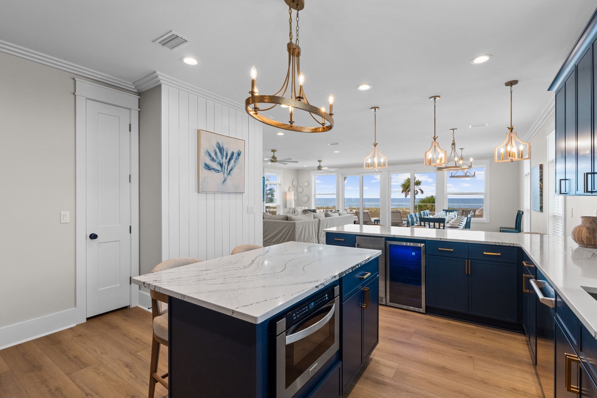 Open kitchen and living space where coastal elegance meets comfort. Rich navy cabinetry contrasts with white marble counters and warm brass fixtures, while ocean views sparkle beyond the windows.