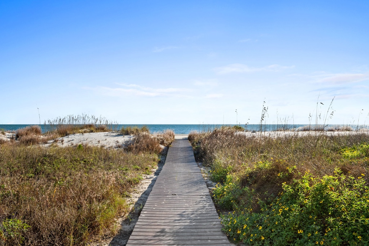 A wooden boardwalk leads through coastal dunes to pristine beach waters under clear blue skies.
