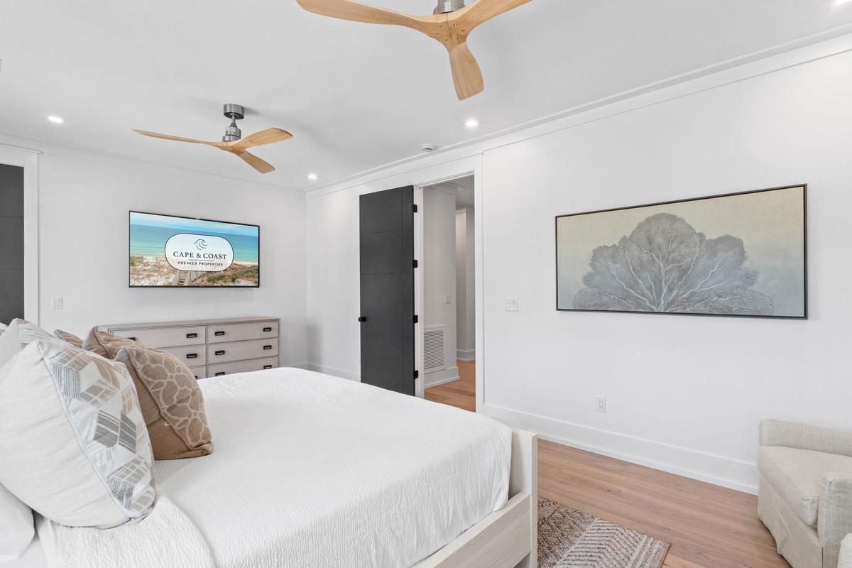 Sink into the plush white bedding and unwind as wooden ceiling fans gently circulate the coastal breeze. Your serene retreat features a smart TV and calming artwork, creating a perfect sanctuary for restful nights.