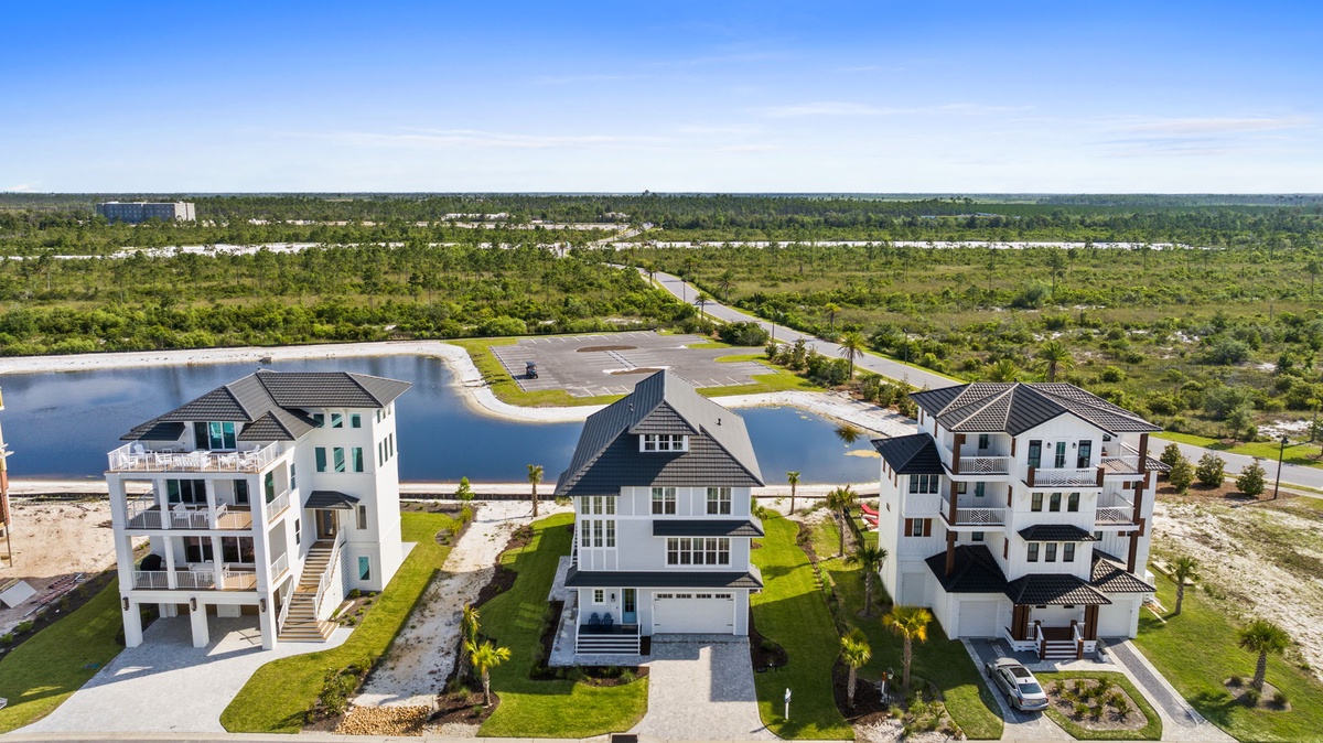 An aerial view of the vacation rental complex, showcasing distinctive coastal architecture nestled along a serene lagoon, surrounded by lush natural landscape and convenient roadway access.