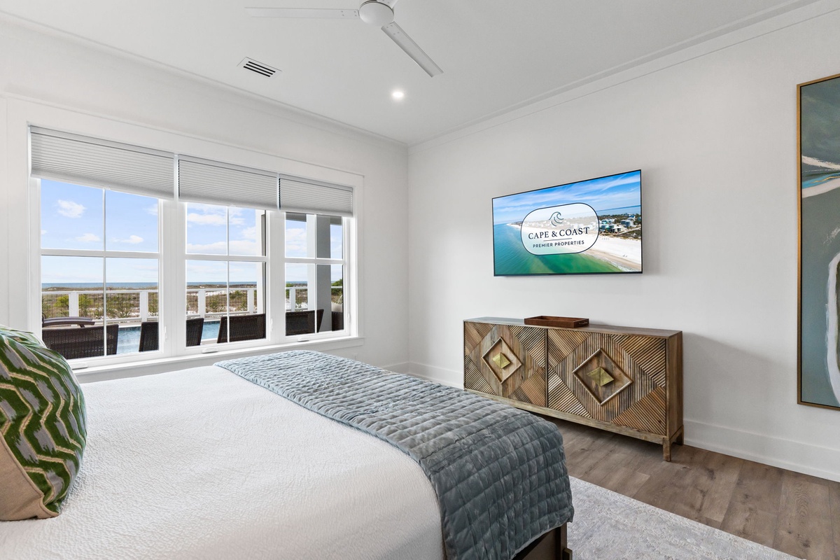 Drift off in your serene bedroom featuring a plush bed, flat-screen TV, and stunning water views framed by elegant blinds.