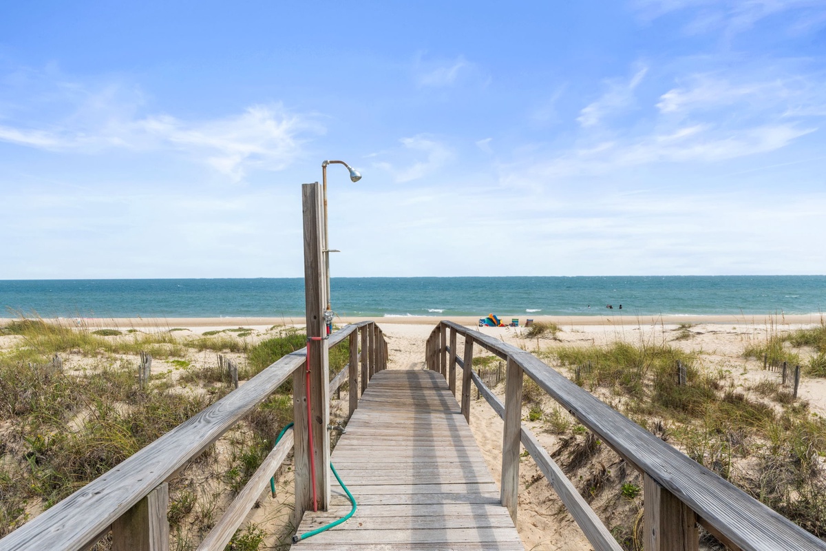 A weathered wooden boardwalk leads across sandy dunes to a pristine beach where calm turquoise waters meet a bright blue sky.