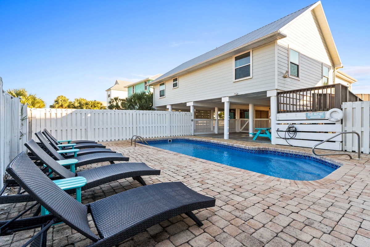 Private pool oasis with comfortable loungers on paved patio, surrounded by white privacy fencing beneath clear blue skies.