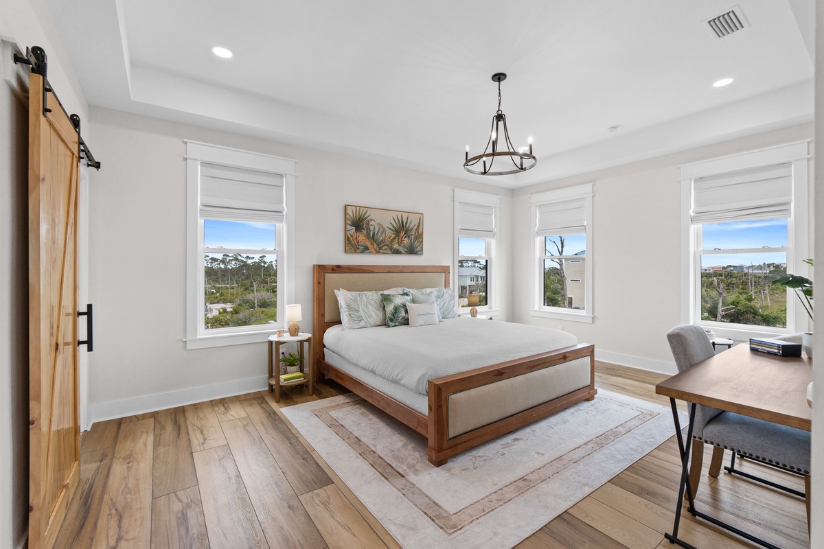 Unwind in your bright, coastal-inspired bedroom featuring a plush king bed, elegant chandelier, and large windows offering serene nature views—perfect for peaceful mornings.