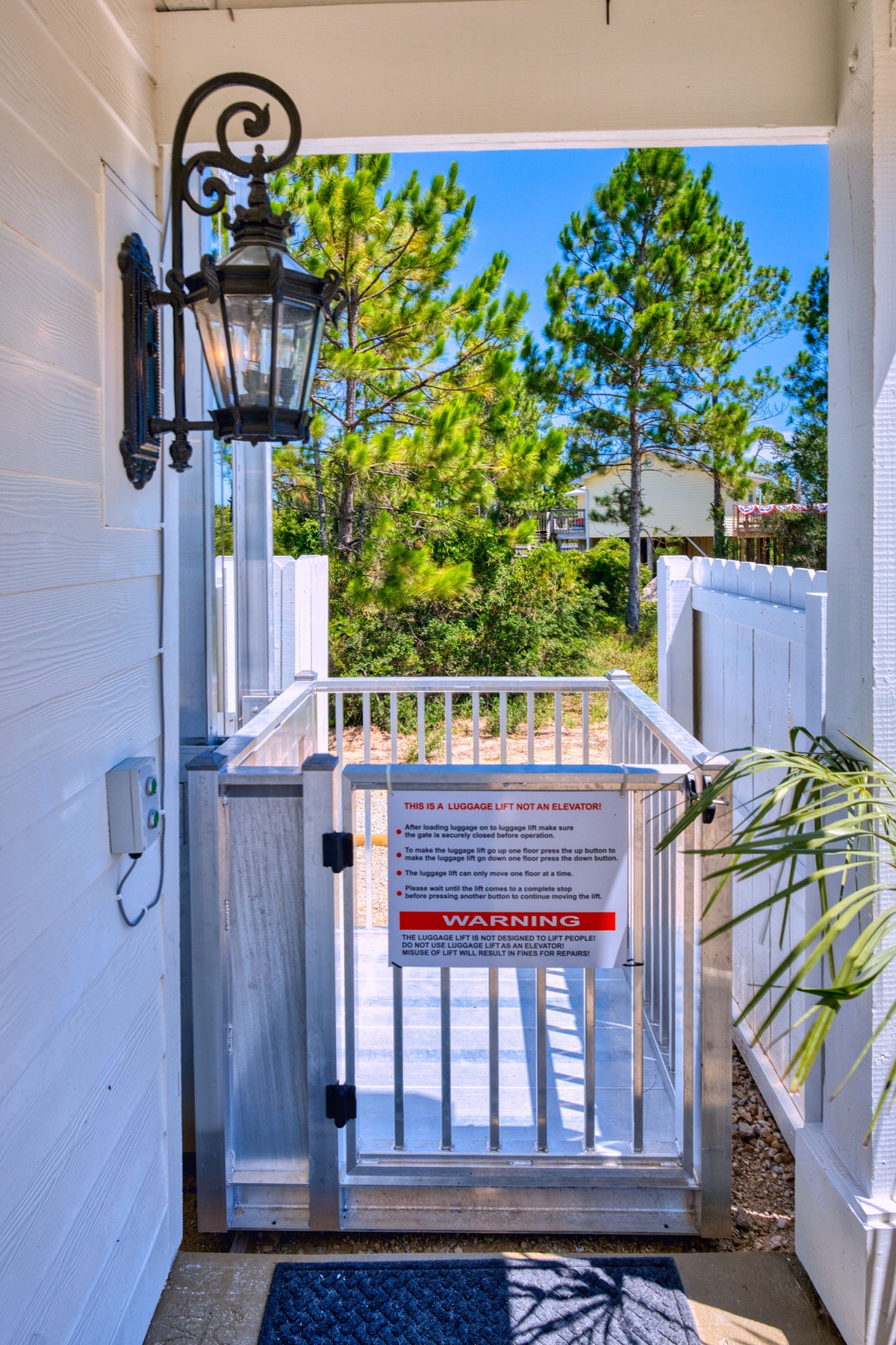 Elevator gate entrance to property, opening to a verdant coastal pine grove and neighboring homes beyond.