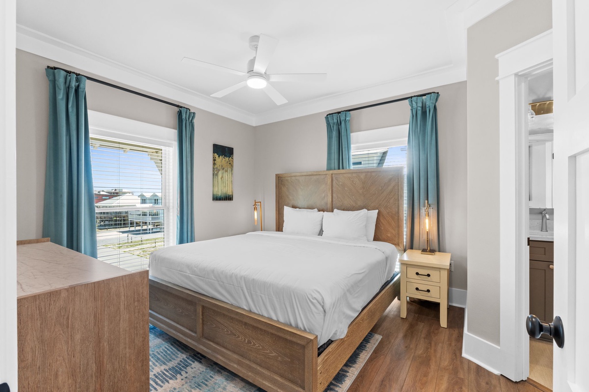 Unwind in your bright, airy bedroom featuring a plush king bed, modern wooden furnishings, ceiling fan, and natural light streaming through teal curtains. Your private en-suite awaits just steps away.