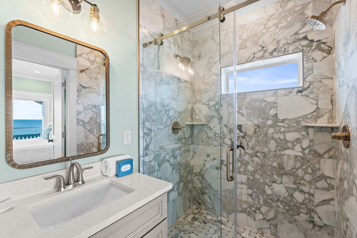 Refresh in your marble-tiled shower with its rainfall showerhead and beach view through the frosted window. The elegant vanity and coastal mirrors add a touch of sophistication to your morning routine.