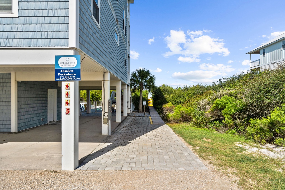 The property entrance features paver walkways bordered by natural coastal vegetation and palm trees, showcasing the beachside setting and sunny skies.