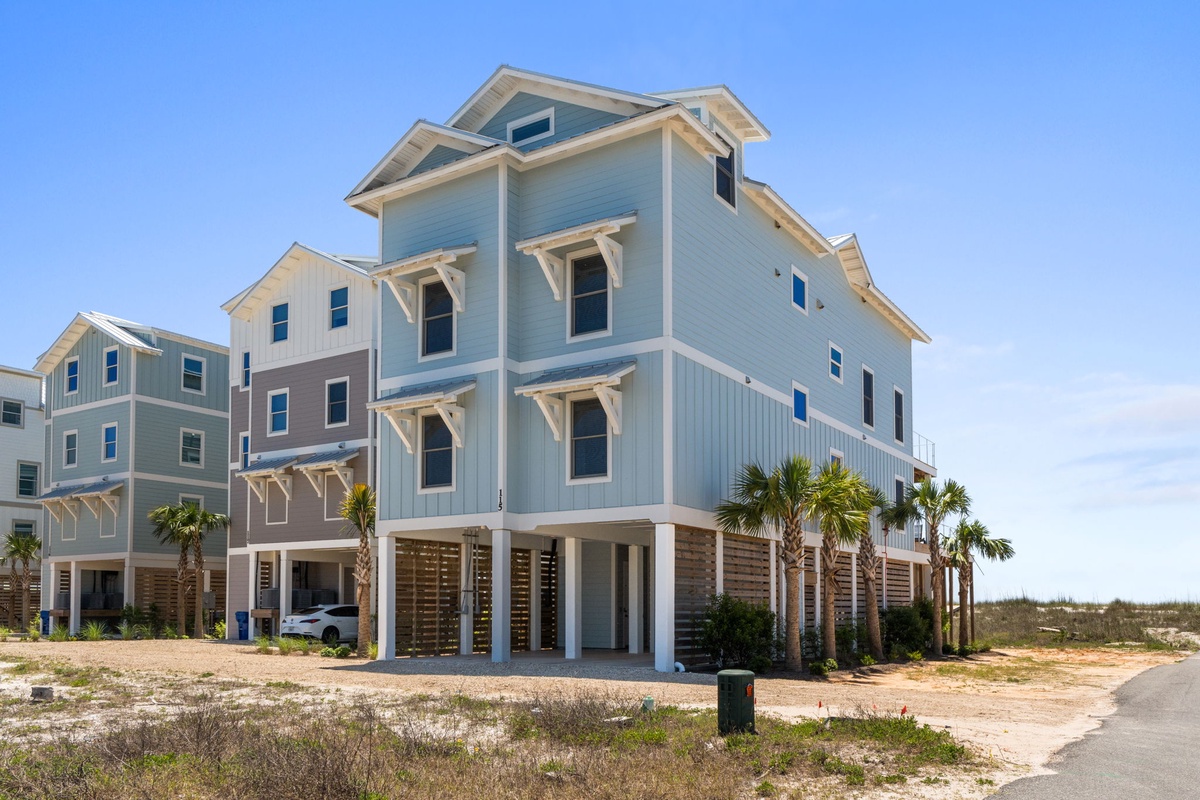 Modern coastal property with elevated design, palm trees, and covered parking situated in a beachside neighborhood with sandy surroundings.
