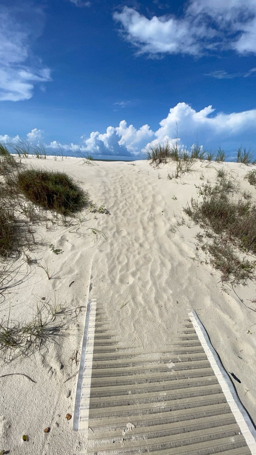 Pristine white sand dunes frame a walkway leading toward the beach, with tufts of natural sea grass dotting the landscape under a vibrant summer sky.