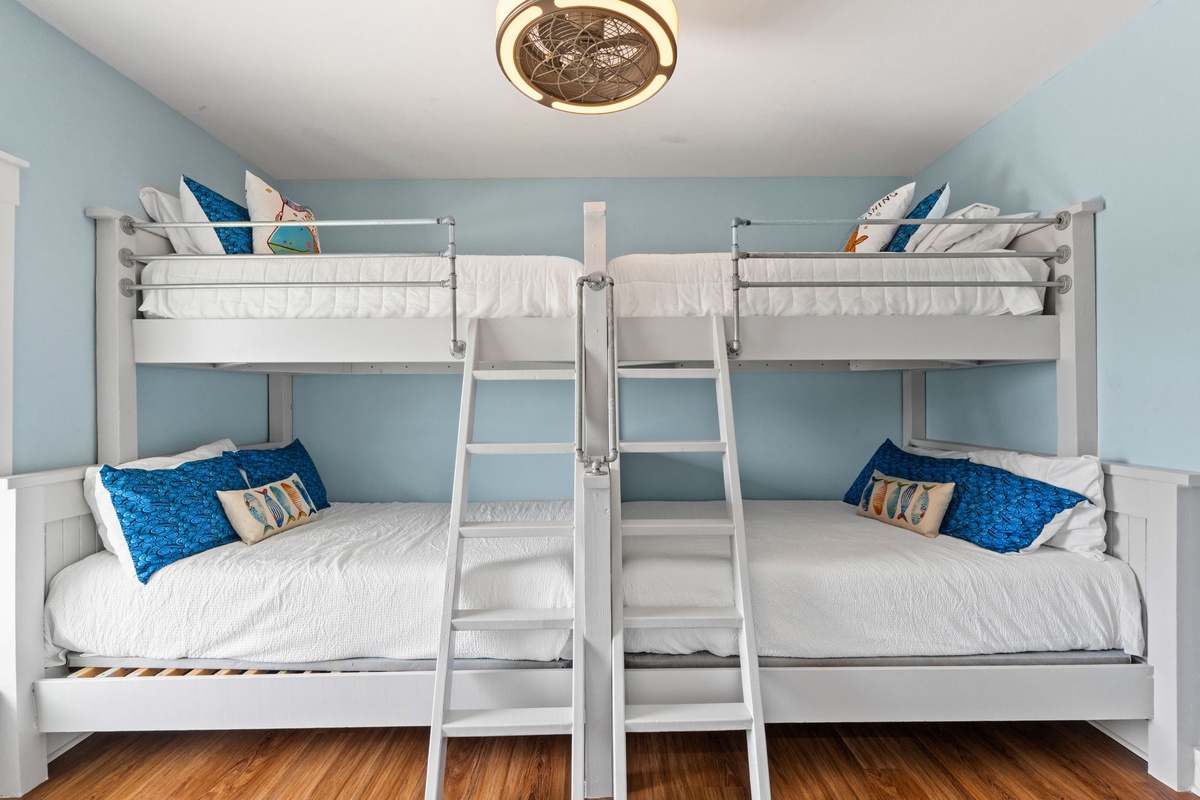 Bright bunk room with coastal charm features twin-over-full beds, crisp white linens with vibrant blue accents, and a playful ceiling fan—perfect for the whole crew.