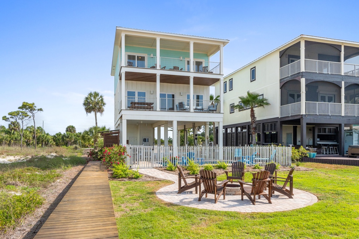 This three-story coastal rental features multiple balconies, covered parking, and a circular fire pit with Adirondack chairs set among lush tropical landscaping and palm trees.