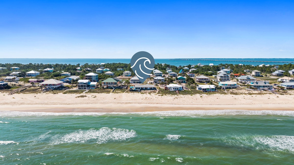 Aerial view of a beachfront community nestled between pristine sandy shores and turquoise waters, showcasing vacation homes amid coastal dunes with a distant bridge spanning the horizon.