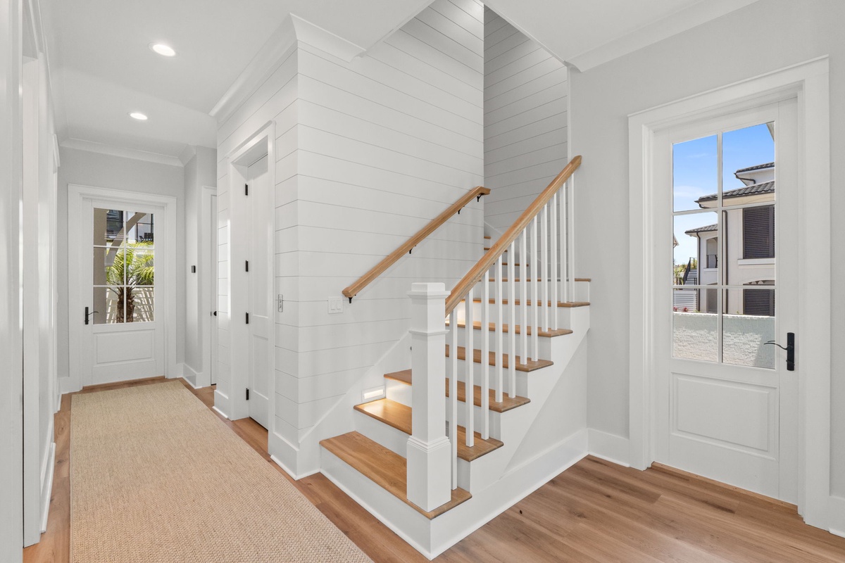 Bright coastal-inspired entryway welcomes you with crisp shiplap walls, natural wood stairs, and views of nearby homes through charming Dutch doors.