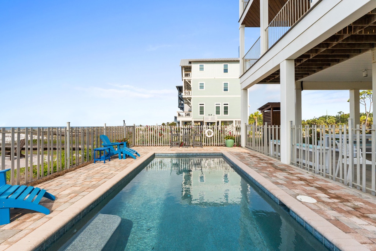 A beachside private pool bordered by brick pavers with bright blue Adirondack chairs and white fencing, overlooking coastal dunes and neighboring beach homes.