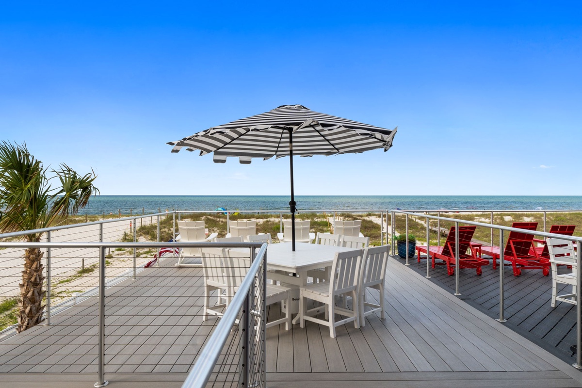 A beachfront deck overlooking turquoise waters with casual white dining furniture and a striped umbrella for your perfect sunny escape.