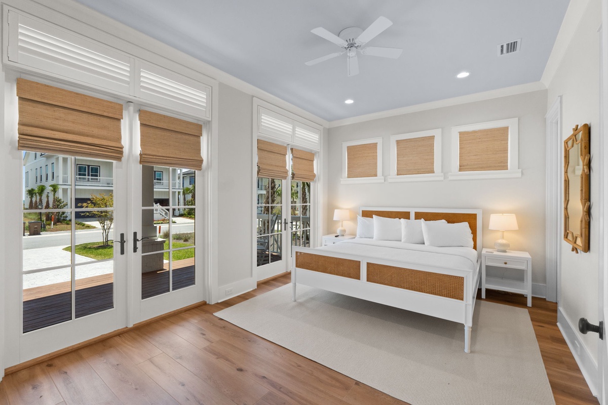 Unwind in your airy bedroom with coastal charm. Natural light pours through French doors, while the ceiling fan keeps you cool. The rattan-accented bed and bamboo shades add warmth.
