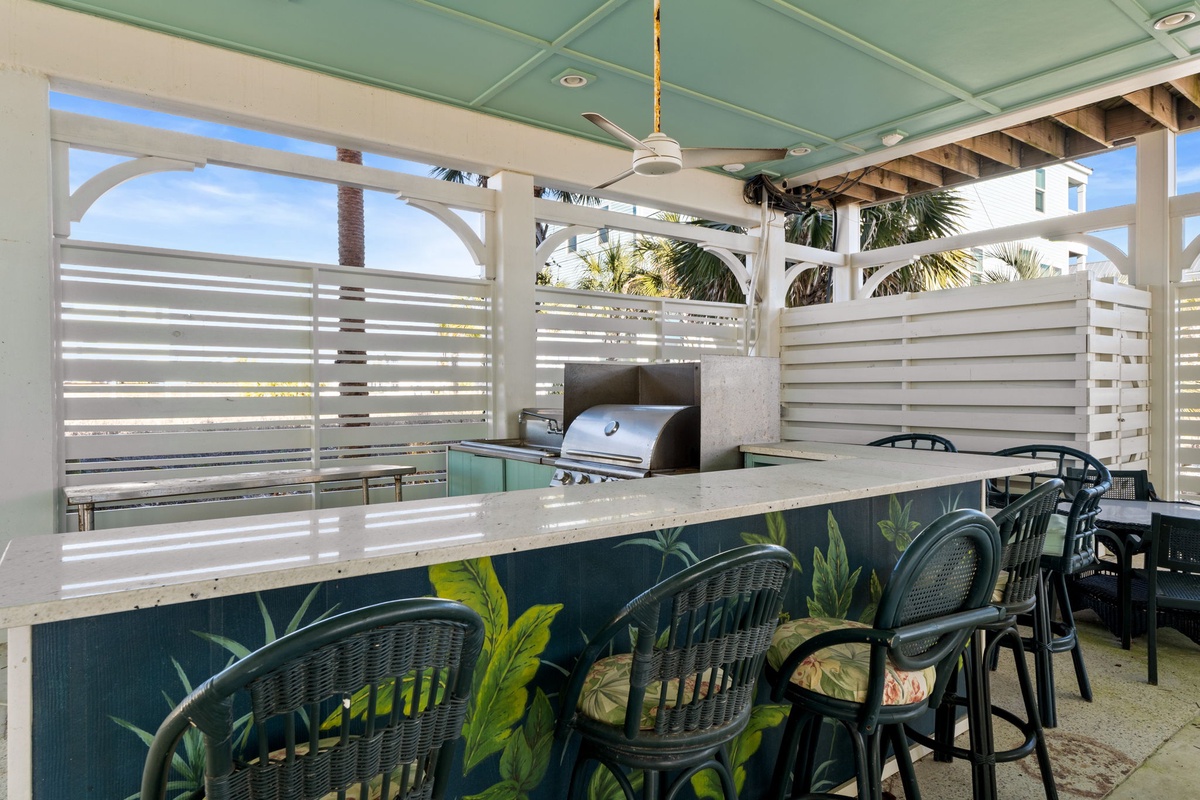 Your covered outdoor kitchen features a built-in grill and breakfast bar seating, perfect for tropical dining with palm tree views.