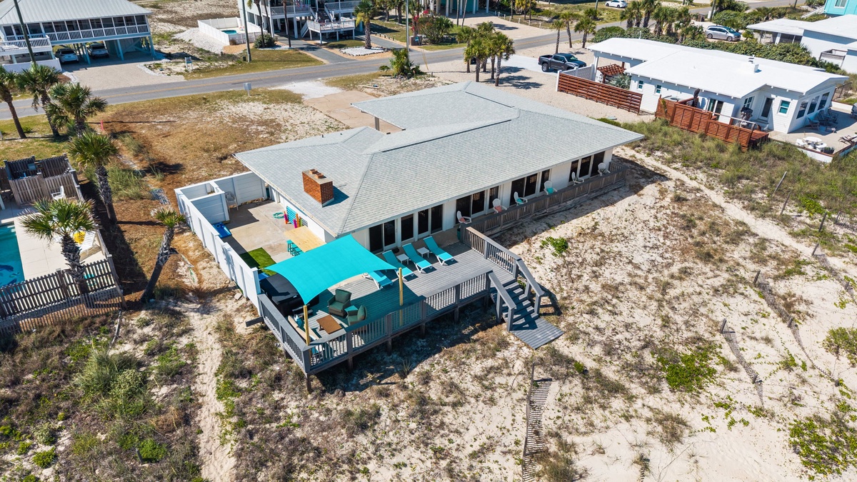 Aerial view showcasing the beachfront property and surrounding coastal neighborhood with palm trees and sandy dunes, located moments from the shore.