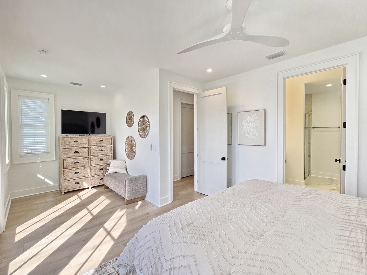 Your bright bedroom features coastal charm with natural wood furniture, wall art, and streaming sunlight creating peaceful patterns.