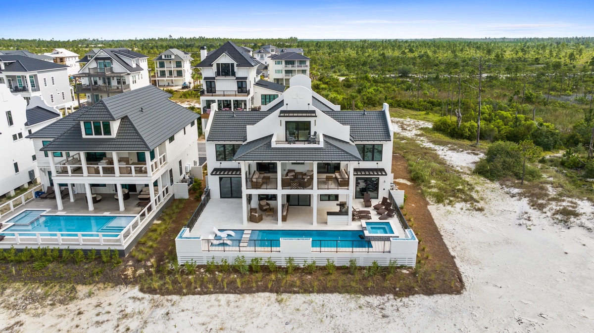 Aerial view of a modern coastal vacation home with dual pools, nestled in a wooded residential area with natural dune landscape and nearby homes.