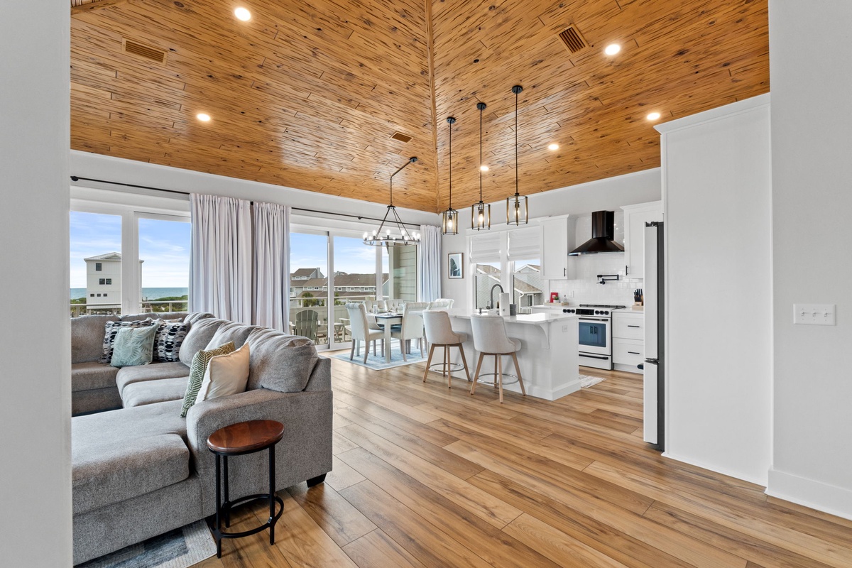 Expansive open-concept living with vaulted wood ceilings, modern kitchen, and waterfront views through floor-to-ceiling windows, blending comfort and elegance for your stay.