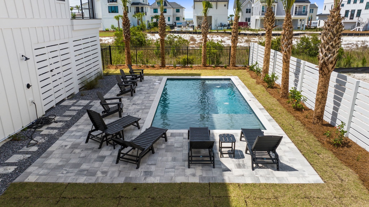 Aerial view of a private pool surrounded by contemporary patio loungers, lush palm landscaping, and privacy fencing in a coastal vacation rental community.