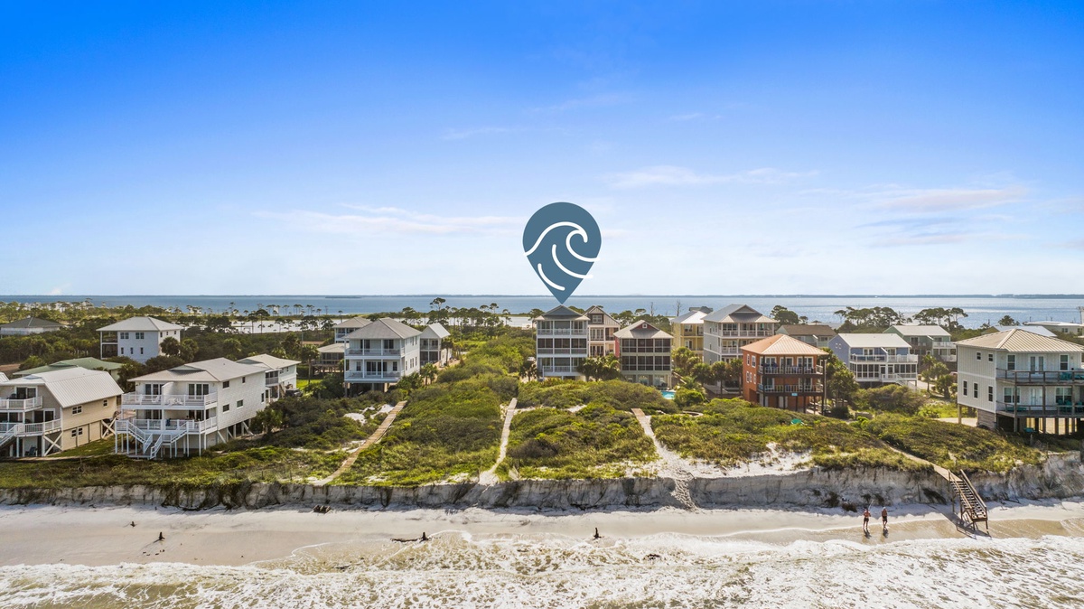 An aerial view captures a coastal community nestled between sandy shores and calm blue waters, with beach access stairs leading down from the dune line.