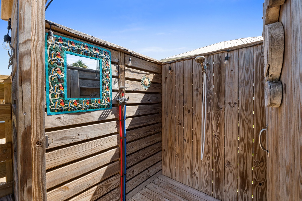 A charming wooden outdoor shower awaits, complete with a colorful decorative mirror and warm sunlight streaming overhead—perfect for rinsing off after beach adventures or soaking up coastal vibes.