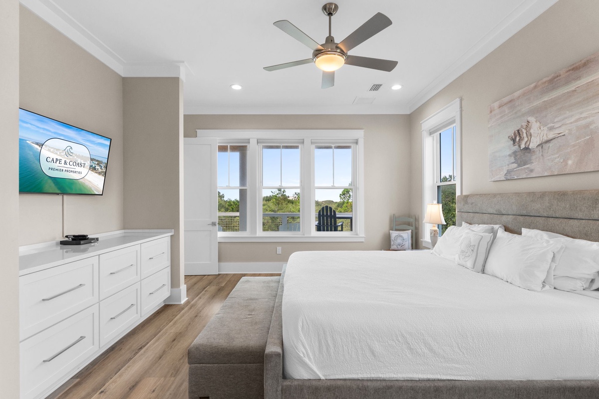 Unwind in your serene bedroom with plush bedding, a ceiling fan for comfort, and windows framing peaceful water views—your coastal retreat awaits.