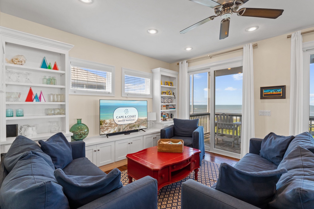 Bright coastal living room with plush seating and floor-to-ceiling windows framing spectacular ocean views. Sliding doors open to your private deck where fresh sea breezes and waves become your everyday soundtrack.
