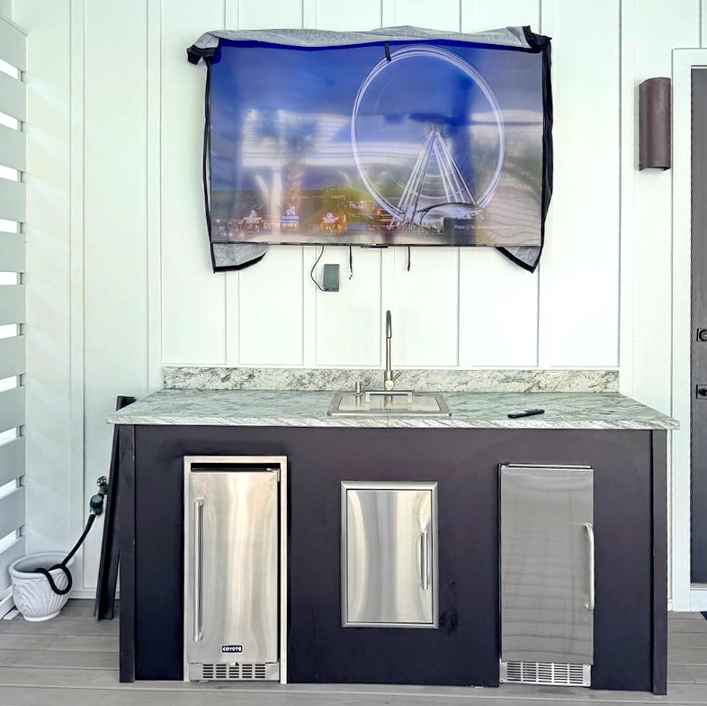 Modern outdoor wet bar features sleek granite countertop, stainless appliances, and sink, perfect for entertaining under the coastal artwork.