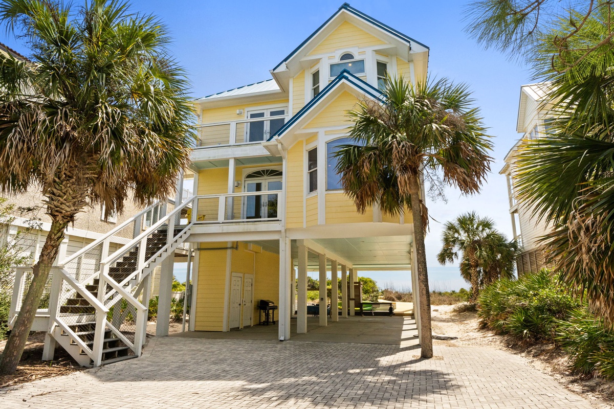 A charming yellow beach house elevated on stilts, surrounded by swaying palm trees and tropical landscaping in a coastal setting.