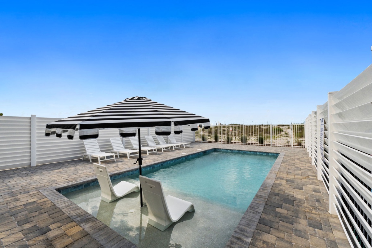 A coastal property featuring a private pool with sun loungers and shaded seating, set against the backdrop of the surrounding beachfront area.