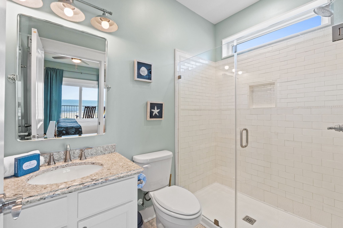 Your private bathroom features a walk-in shower, coastal-inspired decor, and a mirror reflecting the bedroom's ocean view—an inviting retreat after a beach day.